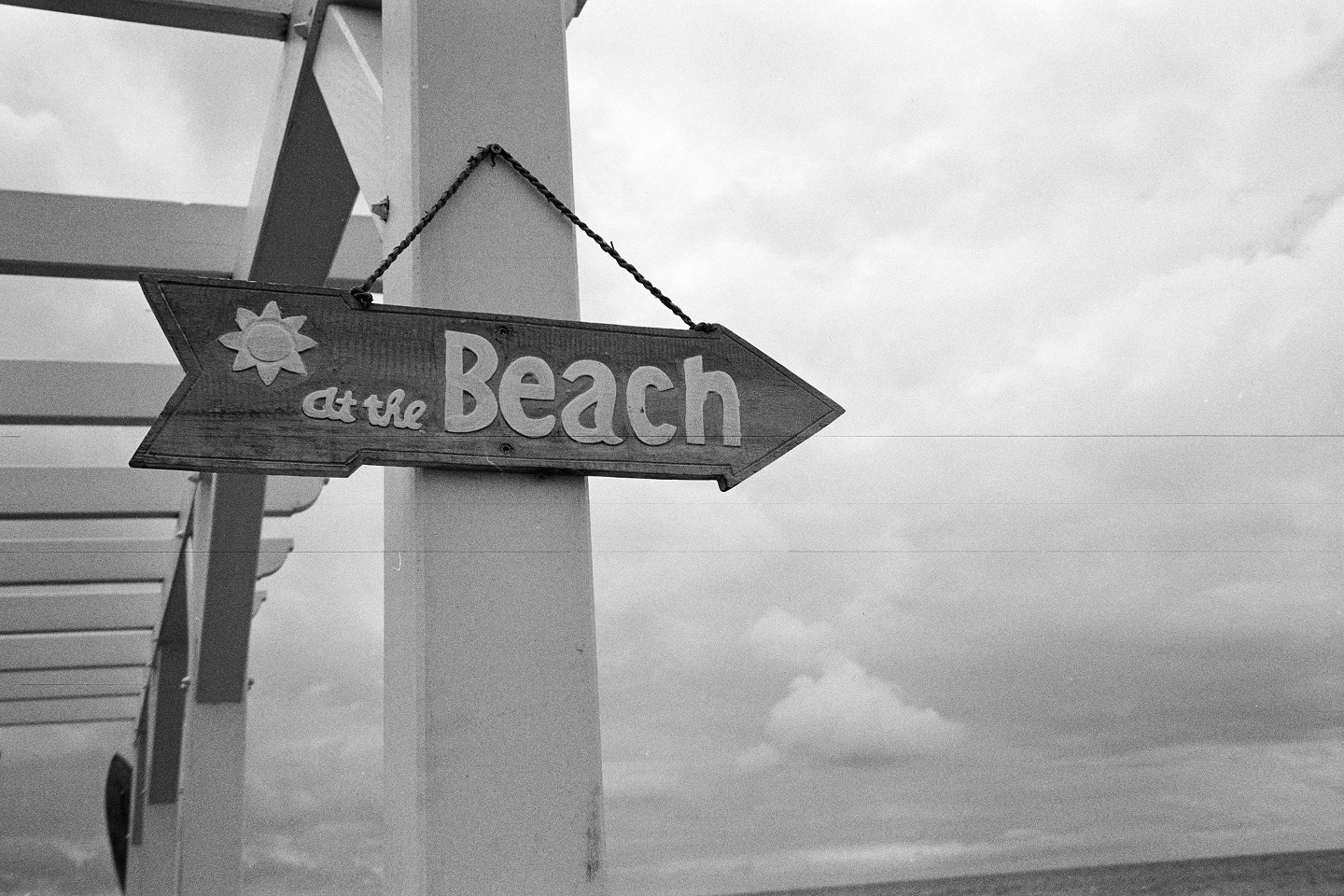 B&W close-up of a rustic wooden arrow sign reading "at the Beach" hanging from a white pergola beam against a cloudy sky.