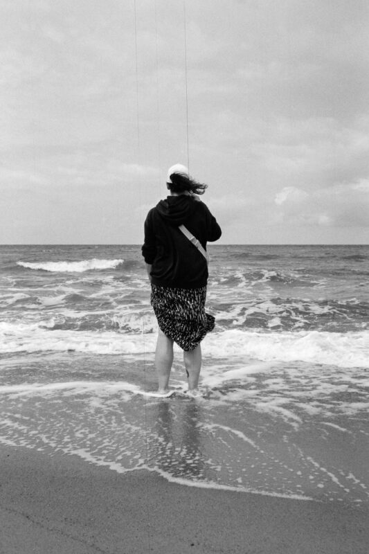 Grainy B&W photo of a person from behind, standing in the shallow surf of the windy sea, wearing a hoodie and a patterned skirt.