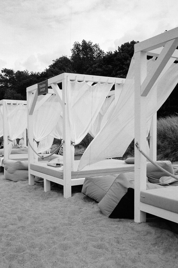 B&W film photo of white luxury beach daybeds with flowing canopy drapes arranged on the sand, with trees in the background.