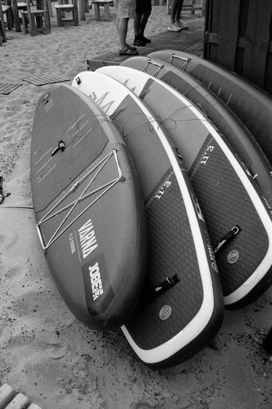 B&W close-up of several stand-up paddleboards stacked on the beach sand, showcasing their textured grip pads and brand names.