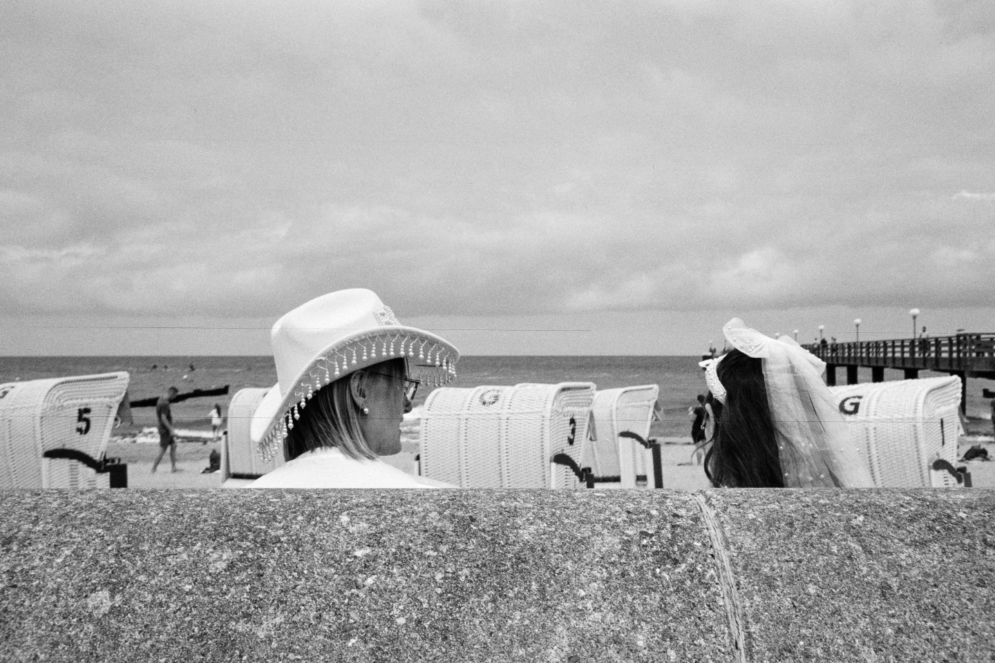 Grainy B&W photo of two women in white decorative hats looking at each other behind a concrete wall, with beach chairs in the back.