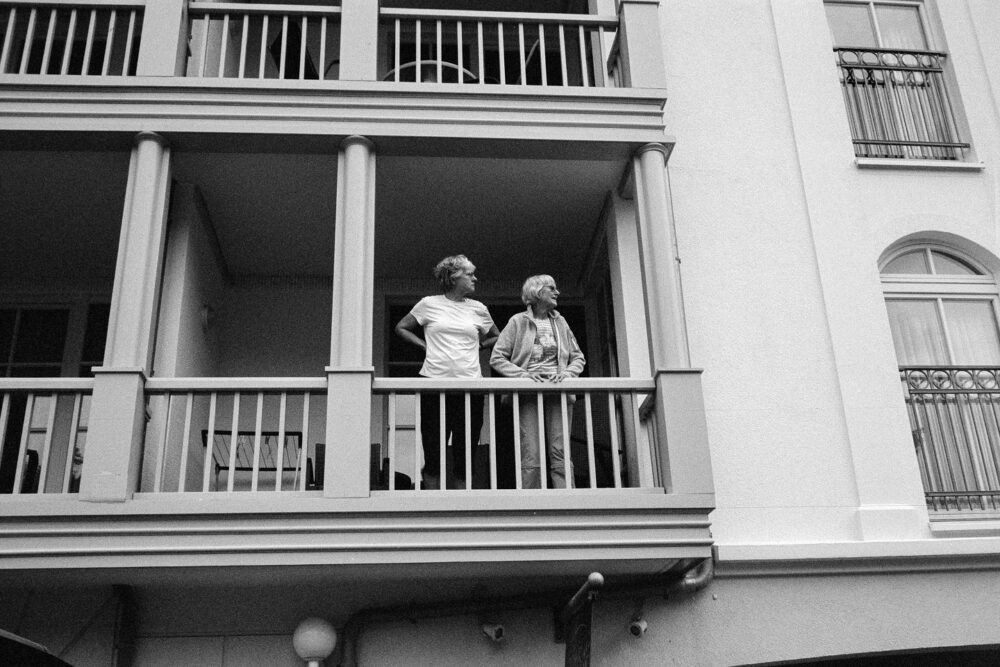 B&W film photo of two women standing on a wooden resort balcony, looking out from a grand white seaside villa facade.