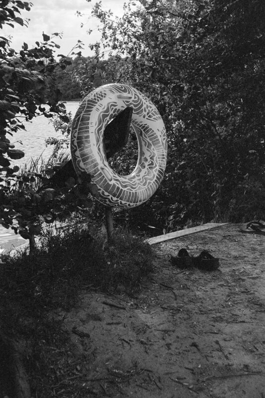 Grainy B&W film photo of a patterned swim ring hung on a post by a lakeshore with sandals on the sandy ground.