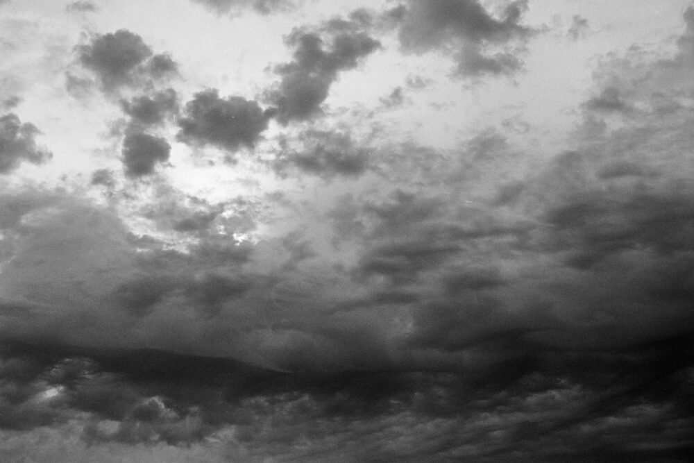 Grainy B&W film photo of a dramatic atmospheric sky filled with swirling dark clouds and soft light breaks.