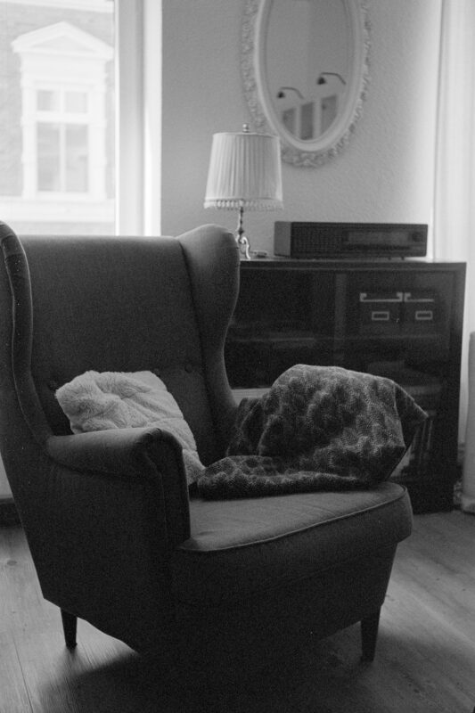 Grainy B&W film photo of a comfortable wingback armchair with a soft blanket in a cozy room with vintage decor.