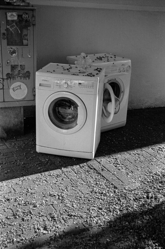 B&W film photo of two abandoned washing machines sitting outdoors on paved ground scattered with tiny debris.
