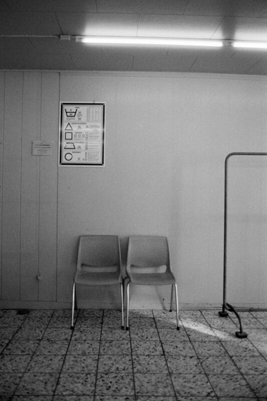 Grainy B&W film photo of two empty chairs in a minimalist waiting area next to a laundry instruction sign and a clothing rack.