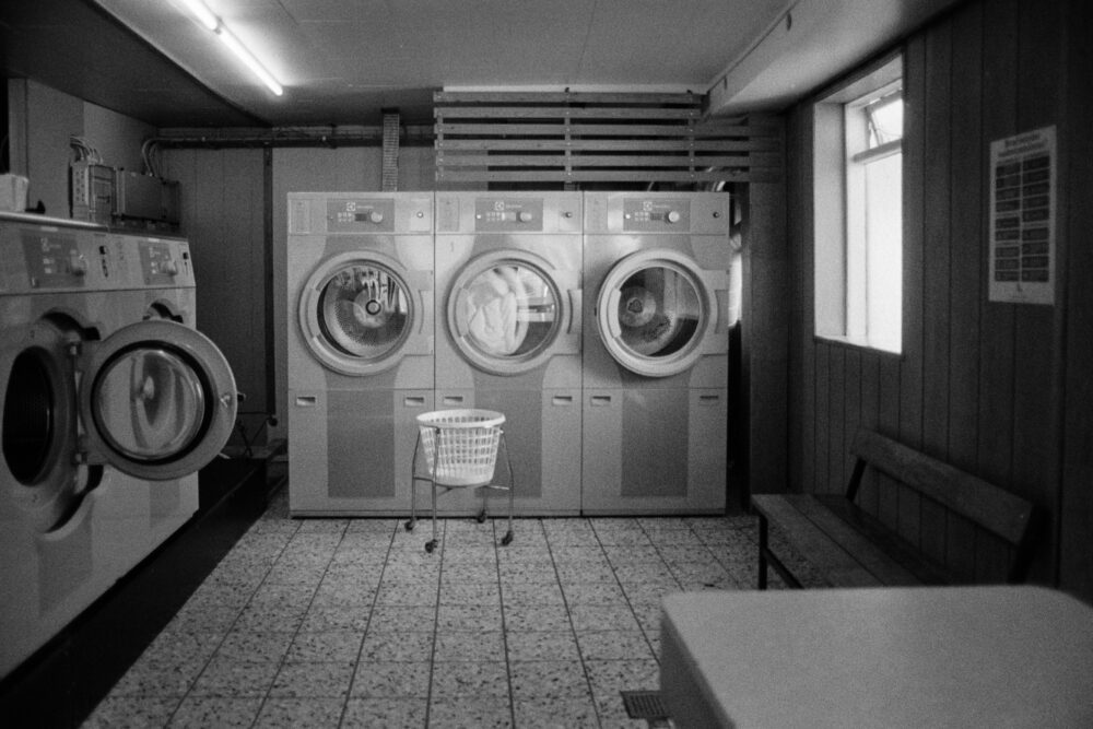 B&W film photo of a retro laundromat interior featuring three aligned washing machines, tiled floors, and a wire laundry basket.