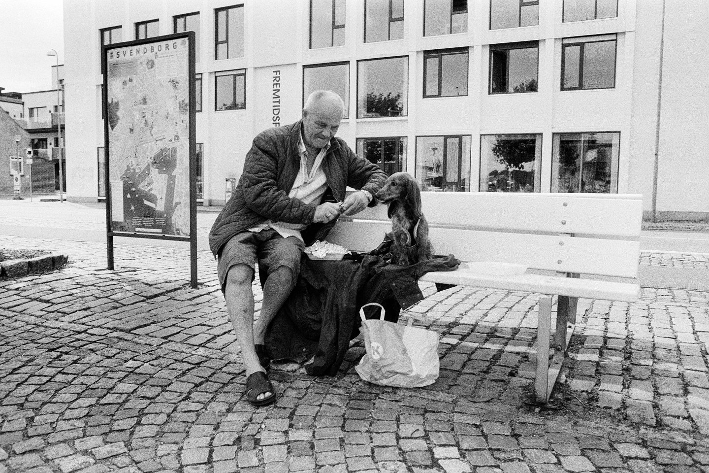 B&W film photo of an older man and his dachshund sharing a meal on a cobblestone street bench near a Svendborg city map sign.