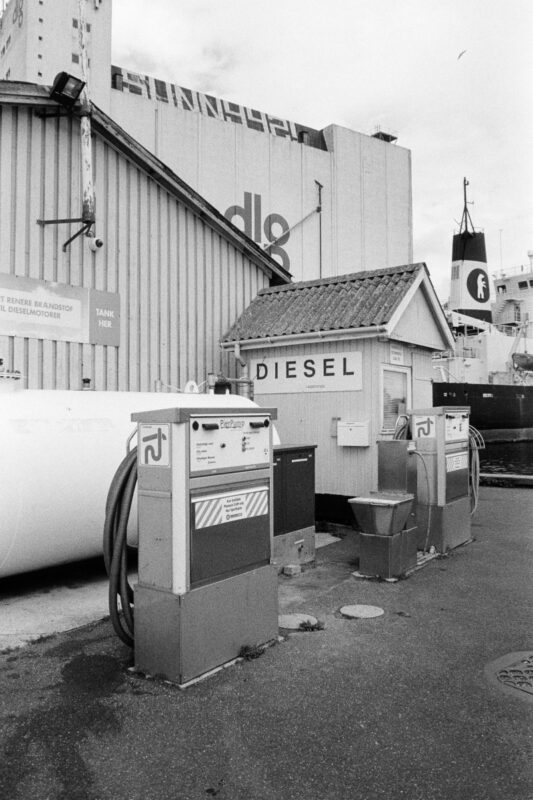 Grainy B&W shot of two vintage marine diesel pumps and a small wooden hut at an industrial harbor, backed by a large silo.
