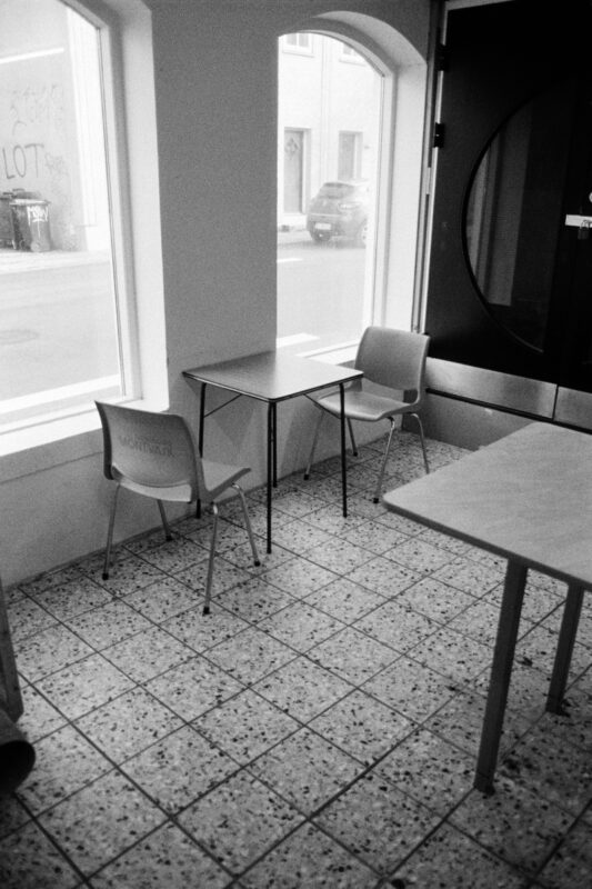 Minimalist B&W film photo of an empty table and two chairs by a large bright window looking out onto a quiet street.
