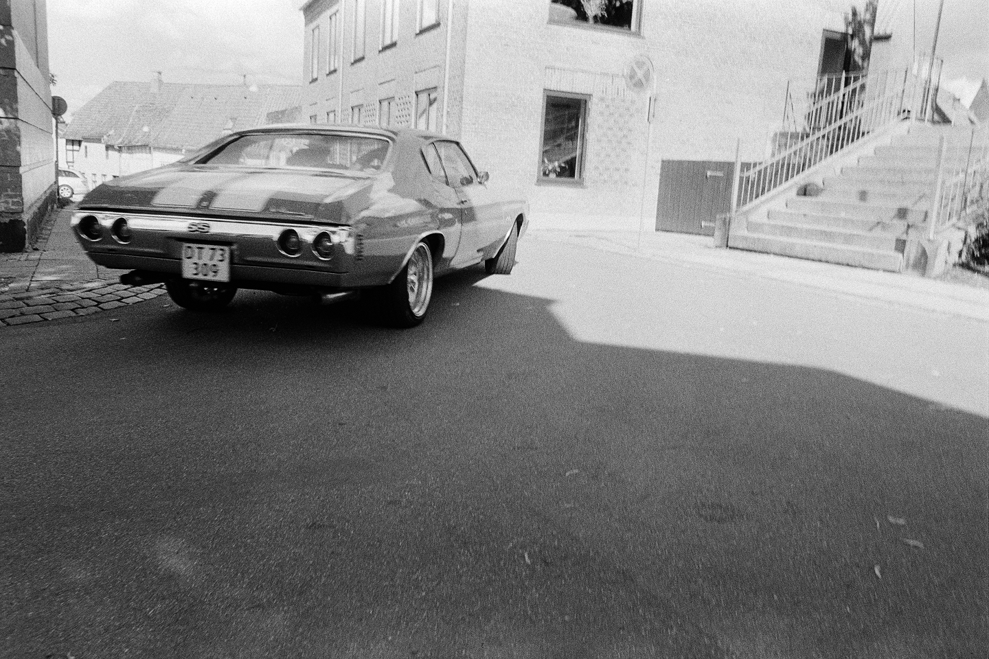 Grainy B&W film photo showing a low-angle rear view of a classic muscle car driving on an empty street next to building stairs.