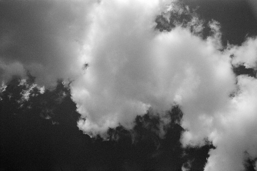 High-contrast B&W film photo looking up at bright, fluffy clouds floating against a dark, textured sky.