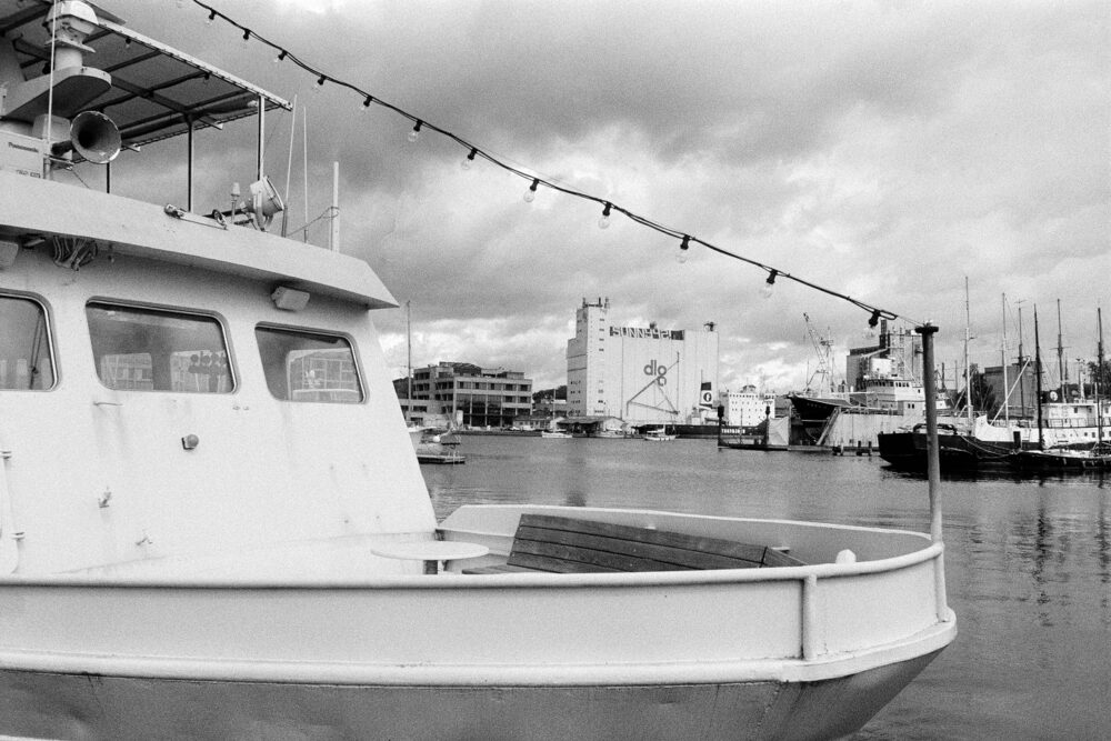 B&W film photo from a boat deck with overhead string lights, looking out over an industrial harbor and a large silo.