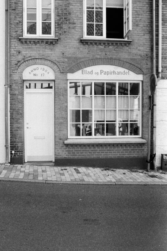 B&W film photo of a brick facade featuring a white door marked "ANNO 1904" and a window sign "Blad og Papirhandel."